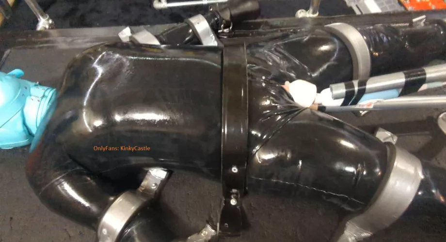 Catsuit girl hooded, mittened, screwed down using metal straps and tek bolts, and then vibed and held in place with a dildo.