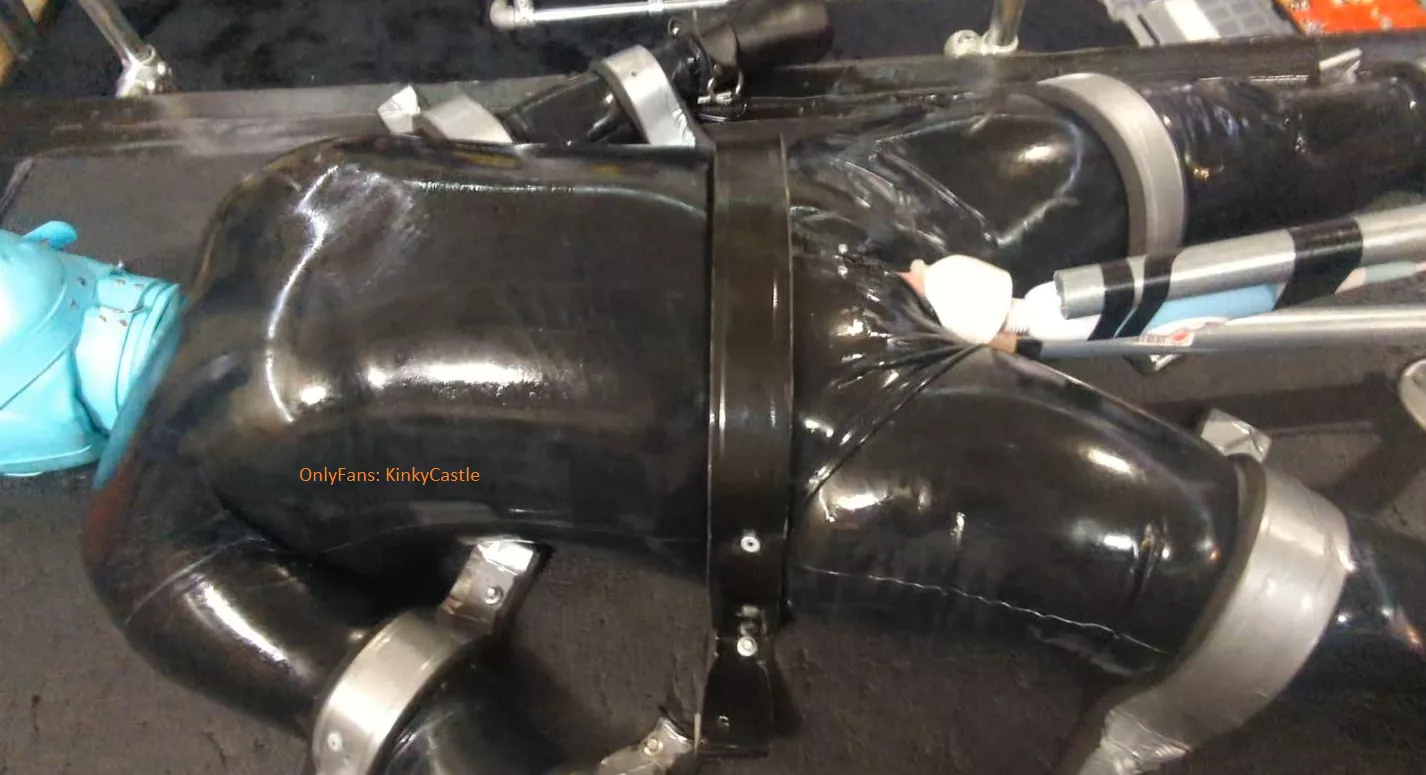 Catsuit girl hooded, mittened, screwed down using metal straps and tek bolts, and then vibed and held in place with a dildo.