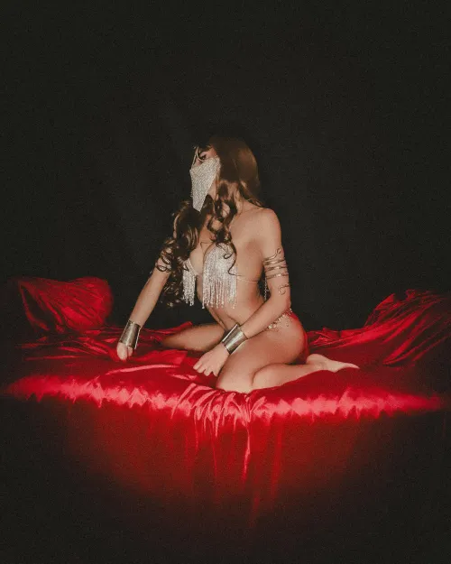 Reclining on rich red fabric, her jeweled mask and soft posture evoke a quiet, luxurious harem fantasy